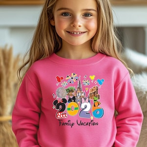 May include: A pink sweatshirt with a colorful graphic that says "2020 Family Vacation." The graphic includes images of Mickey and Minnie Mouse, a castle, and other vacation-themed icons. The sweatshirt is worn by a young person.