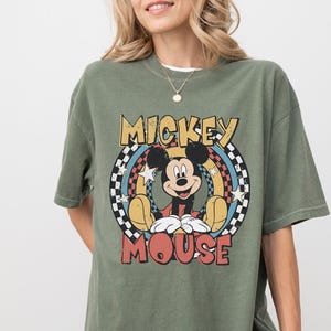 Puede incluir: Una camiseta de manga corta color verde oliva con un gr&aacute;fico de Mickey Mouse centrado en un marco circular. El dise&ntilde;o incluye un borde a cuadros blanco y negro, detalles en amarillo y azul, estrellas blancas y el texto MICKEY MOUSE en rojo.