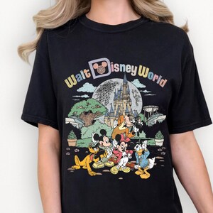 May include: Black t-shirt featuring a colorful graphic of Disney characters, including Mickey Mouse, Minnie Mouse, Donald Duck, and Goofy, in front of a castle and the words "Walt Disney World". The shirt has a crew neck and short sleeves.