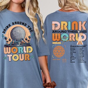 May include: Comfort blue jean t-shirt with a colorful graphic. The front features the text "DRINK AROUND THE WORLD TOUR 26" with a globe design. The back has "DRINK AROUND THE WORLD" and a list of countries.