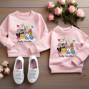 May include: Two pink sweatshirts with a colorful graphic featuring Mickey and Minnie Mouse, the number 2020, and the words "Family Vacation." White sneakers are at the bottom. A bouquet of pink roses rests on a wooden surface.