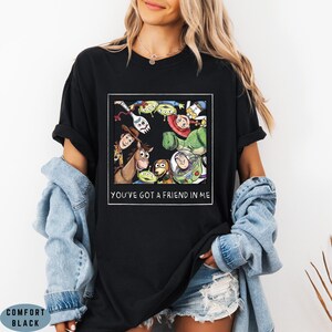 May include: A black short-sleeved t-shirt featuring a square graphic print of various animated characters arranged in a circle. Below the characters, the text reads YOU'VE GOT A FRIEND IN ME. The shirt is worn with a light blue denim jacket and denim shorts.