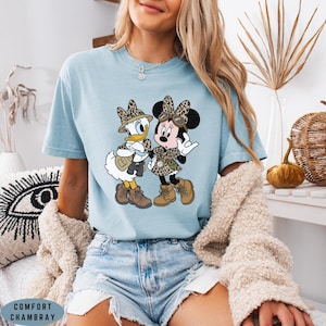 K&ouml;nnte beinhalten: Ein hellblaues T-Shirt mit einem Grafikdruck von Daisy Duck und Minnie Mouse, die in Safari-Kleidung mit Leopardenmuster-Akzenten gekleidet sind, einschlie&szlig;lich H&uuml;ten, Westen und Stiefeln. Das T-Shirt ist als Comfort Chambray gekennzeichnet.