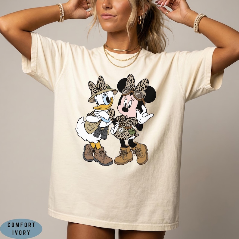 K&ouml;nnte beinhalten: Ein elfenbeinfarbenes Kurzarm-T-Shirt mit einem Grafikdruck von Daisy Duck und Minnie Mouse in Safari-Outfits mit Leopardenmuster, H&uuml;ten und Stiefeln. Die Figuren sind mit Fernglas und Kompass dargestellt. Der Text COMFORT IVORY ist zu sehen.