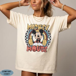 Puede incluir: Una camiseta de color marfil con un gr&aacute;fico de Mickey Mouse centrado en un marco circular. El dise&ntilde;o incluye un borde a cuadros, estrellas y el texto MICKEY MOUSE en letras negritas y desgastadas. La camiseta est&aacute; etiquetada como Comfort Ivory.