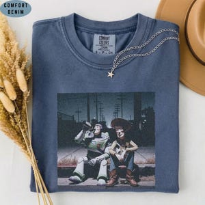 May include: A blue crew neck t-shirt featuring a graphic print of two animated characters sitting together at night. The shirt has a Comfort Colors label at the inner neckline and is accented with a delicate silver chain necklace with a star charm.
