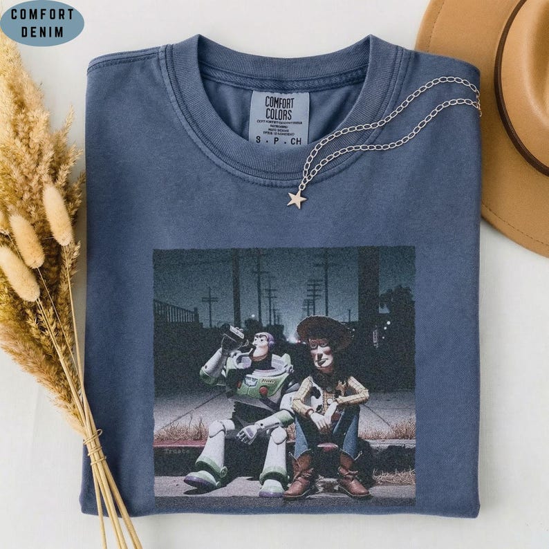 May include: A blue crew neck t-shirt featuring a graphic print of two animated characters sitting together at night. The shirt has a Comfort Colors label at the inner neckline and is accented with a delicate silver chain necklace with a star charm.