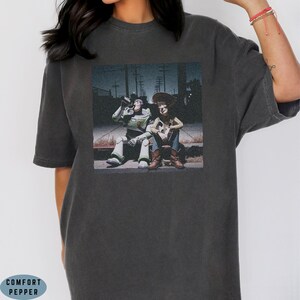 May include: A charcoal gray short-sleeve t-shirt featuring a graphic print of the characters Buzz Lightyear and Woody sitting together on a curb at night. The shirt has a crew neckline and a relaxed fit, showcasing a detailed cinematic scene.