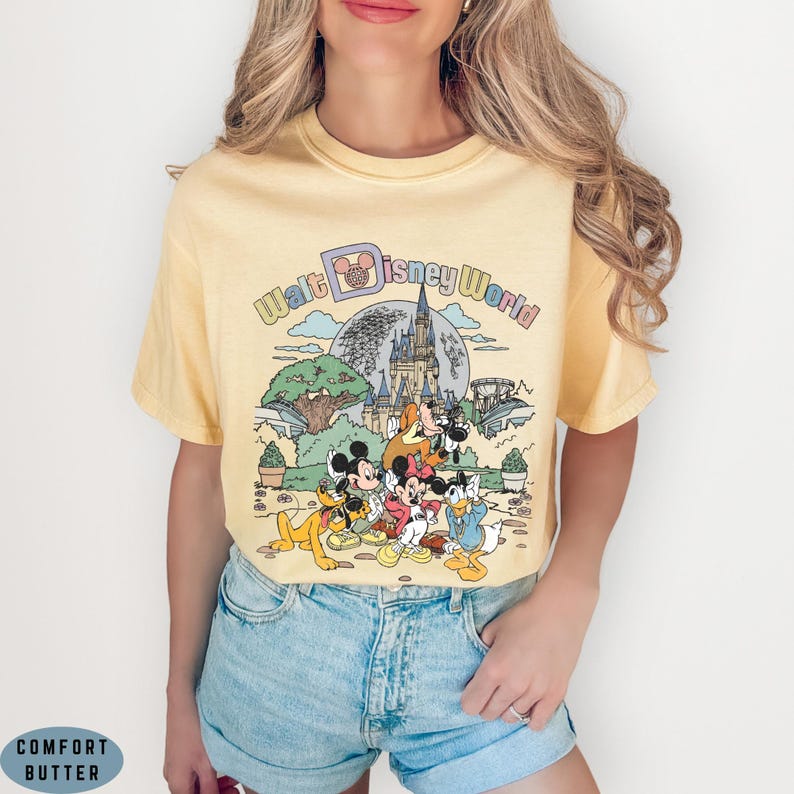 May include: Yellow t-shirt featuring a colorful graphic of Disney characters, including Mickey Mouse, Minnie Mouse, and Donald Duck, in front of a castle and the words "Walt Disney World". The shirt is a light yellow color.