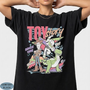 May include: A black t-shirt featuring a graphic print of Woody, Buzz Lightyear, and Rex from Toy Story. The design includes the text TOY story, SINCE 1995, Woody, Buzz, and Rex in a colourful, retro-inspired style with geometric shapes and brush strokes.