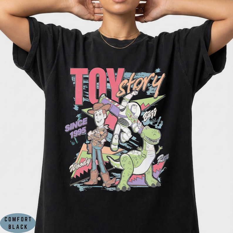 May include: A black t-shirt featuring a graphic print of Woody, Buzz Lightyear, and Rex from Toy Story. The design includes the text TOY story, SINCE 1995, Woody, Buzz, and Rex in a colourful, retro-inspired style with geometric shapes and brush strokes.