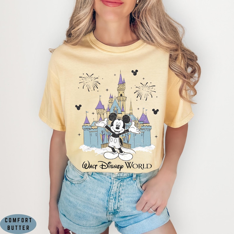 Op de afbeelding: Lichtgeel T-shirt met een afbeelding van Mickey Mouse voor het kasteel van Assepoester in Walt Disney World. Het ontwerp bevat vuurwerk en Mickey Mouse-iconen. De tekst "Walt Disney World" staat eronder.
