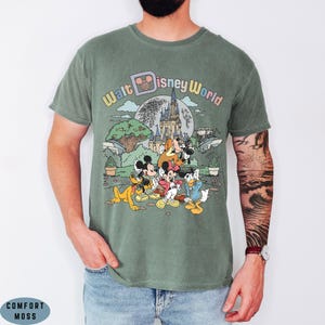 May include: Sage green t-shirt featuring a colorful graphic of Disney characters, including Mickey Mouse, Minnie Mouse, and Donald Duck, in front of Cinderella's castle. The text "Walt Disney World" is arched above the scene.