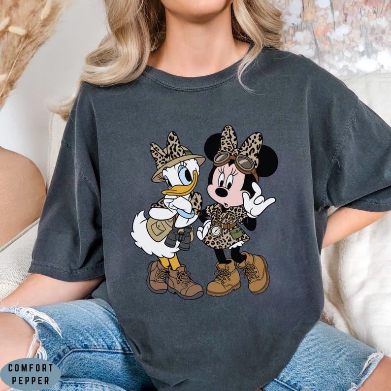 K&ouml;nnte beinhalten: Ein anthrazitfarbenes Kurzarm-T-Shirt mit einem Grafikdruck von Daisy Duck und Minnie Mouse in Safari-Ausr&uuml;stung. Beide Figuren tragen H&uuml;te und Outfits mit Leopardenmuster, braune Stiefel und f&uuml;hren Entdecker-Accessoires wie ein Fernglas mit.