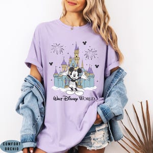 Op de afbeelding: Lichtpaarse t-shirt met een afbeelding van Mickey Mouse voor een kasteel, vuurwerk en de tekst "Walt Disney World". Het shirt is gemaakt van een zacht materiaal.