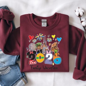 May include: A maroon sweatshirt with a colorful graphic design. The design includes the numbers "2026" with Disney characters and landmarks, and the words "Family Vacation". The sweatshirt is folded on a white surface.