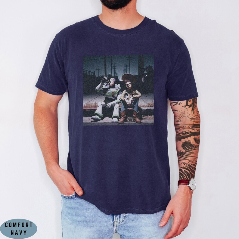 May include: A navy blue short-sleeve t-shirt featuring a graphic print of two toy characters sitting on a curb at night. The design shows a space ranger figure and a cowboy figure in a dark, outdoor setting. The shirt has a crew neckline.