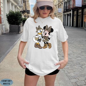 K&ouml;nnte beinhalten: Ein wei&szlig;es Kurzarm-T-Shirt mit einem Grafikdruck von Daisy Duck und Minnie Mouse in Safari-Kleidung, bestehend aus H&uuml;ten und Westen mit Leopardenmuster sowie braunen Stiefeln. Die Figuren stehen im Cartoon-Stil nebeneinander.