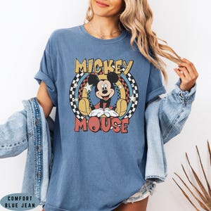Puede incluir: Una camiseta azul de manga corta con un gr&aacute;fico de Mickey Mouse centrado dentro de un patr&oacute;n de cuadros circular. El texto MICKEY MOUSE est&aacute; impreso en amarillo y rojo. La camiseta se lleva debajo de una chaqueta vaquera azul con botones visibles.