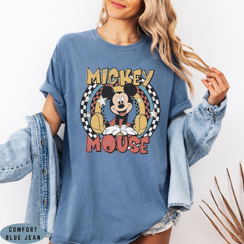 Puede incluir: Una camiseta azul de manga corta con un gr&aacute;fico de Mickey Mouse centrado dentro de un patr&oacute;n de cuadros circular. El texto MICKEY MOUSE est&aacute; impreso en amarillo y rojo. La camiseta se lleva debajo de una chaqueta vaquera azul con botones visibles.