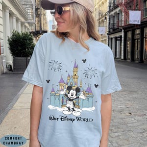 Op de afbeelding: Lichtblauw T-shirt met een afbeelding van Mickey Mouse voor een kasteel en vuurwerk. De tekst "Walt Disney World" staat eronder. Het shirt is gemaakt van Comfort Chambray.