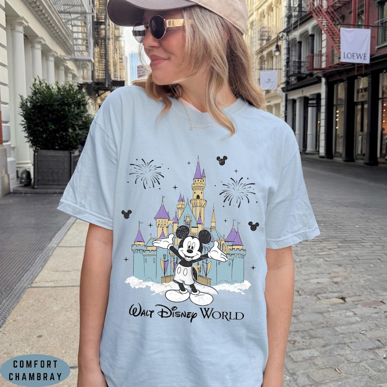 Op de afbeelding: Lichtblauw T-shirt met een afbeelding van Mickey Mouse voor een kasteel en vuurwerk. De tekst "Walt Disney World" staat eronder. Het shirt is gemaakt van Comfort Chambray.