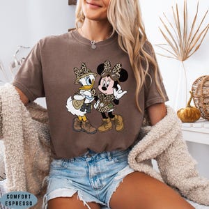 K&ouml;nnte beinhalten: Ein braunes Kurzarm-T-Shirt mit einem Grafikdruck von Daisy Duck und Minnie Mouse, die Safari-Kleidung mit Leopardenmuster, H&uuml;ten und Stiefeln tragen. Das T-Shirt ist mit Comfort Espresso beschriftet.