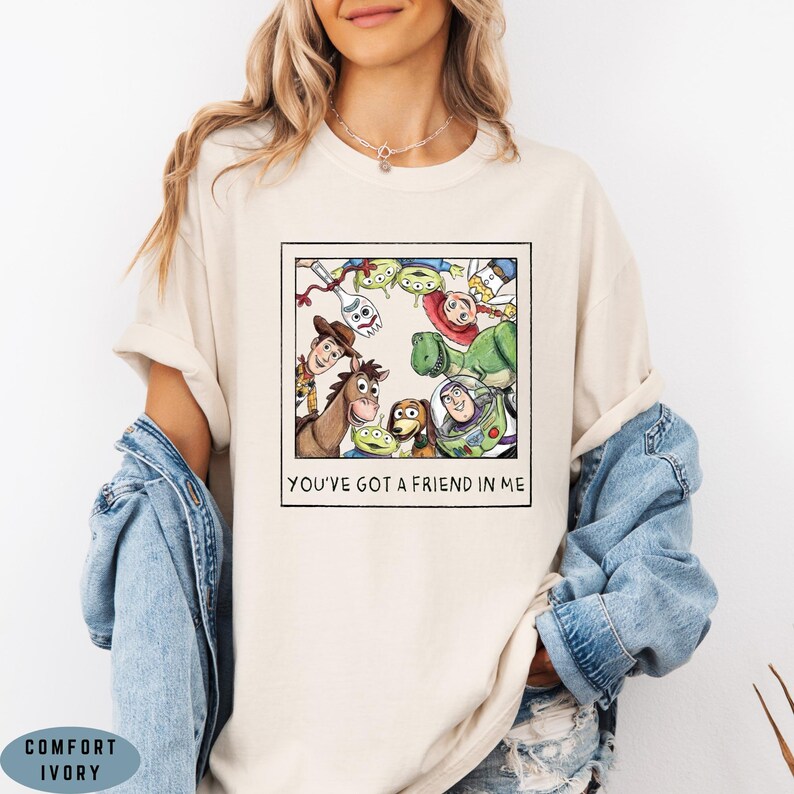 May include: An ivory t-shirt featuring a square graphic print of various Toy Story characters arranged in a circle. The characters include Woody, Buzz Lightyear, Rex, Slinky Dog, Jessie, Forky, and Aliens. Below the graphic is the text YOU'VE GOT A FRIEND IN ME.