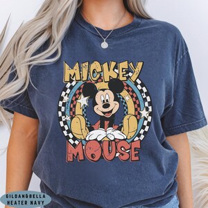 Puede incluir: Una camiseta de manga corta azul marino con un estampado gr&aacute;fico de Mickey Mouse centrado en un dise&ntilde;o circular. El gr&aacute;fico incluye un borde a cuadros en blanco y negro, estrellas amarillas y el texto MICKEY MOUSE en letras negritas.