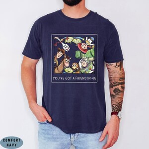 May include: A navy blue t-shirt featuring a square graphic print of various animated characters from Toy Story arranged in a circle. Below the characters, the text reads YOU'VE GOT A FRIEND IN ME in white capital letters. The shirt has short sleeves.