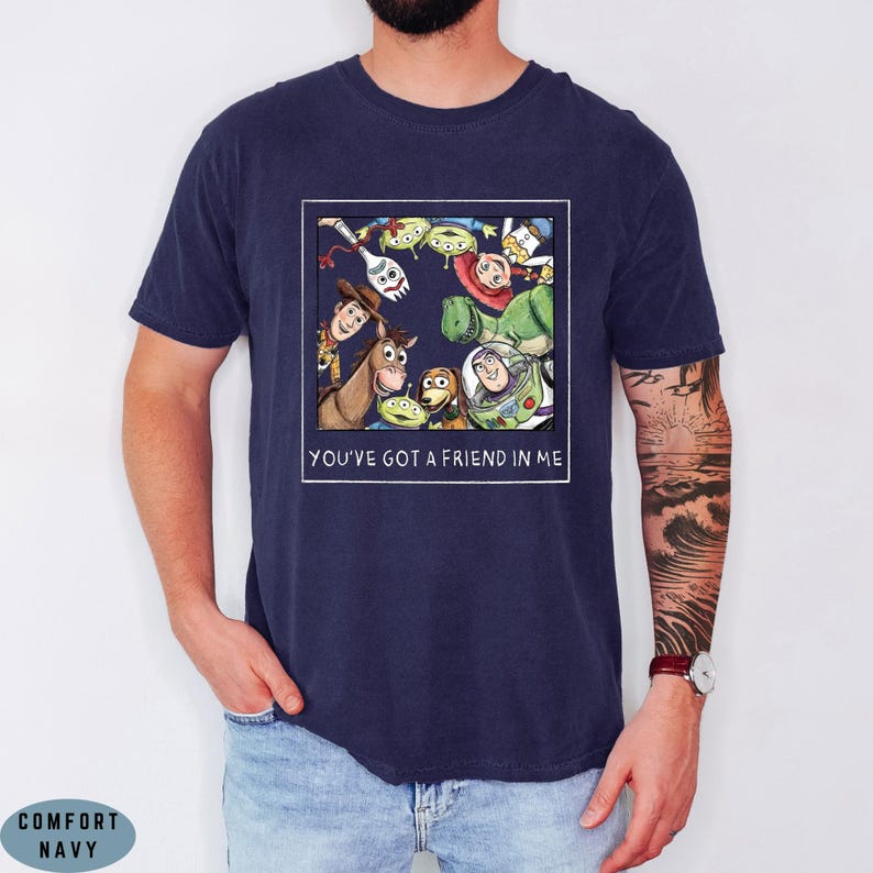 May include: A navy blue t-shirt featuring a square graphic print of various animated characters from Toy Story arranged in a circle. Below the characters, the text reads YOU'VE GOT A FRIEND IN ME in white capital letters. The shirt has short sleeves.