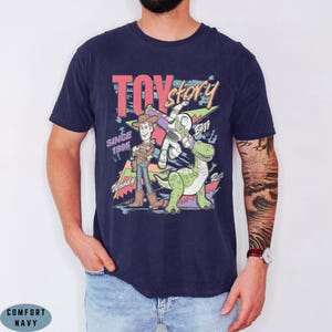 May include: A navy blue short-sleeve t-shirt featuring a graphic print with the text TOY story, SINCE 1995, Woody, Buzz, and Rex. The design includes colourful geometric shapes and outlines of the characters in a retro, illustrated style.