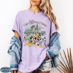 May include: A lilac-colored t-shirt featuring a graphic of Disney characters in front of a castle and the words "Walt Disney World". The design includes Mickey Mouse, Minnie Mouse, Donald Duck, and Goofy. The shirt has short sleeves and a crew neck.