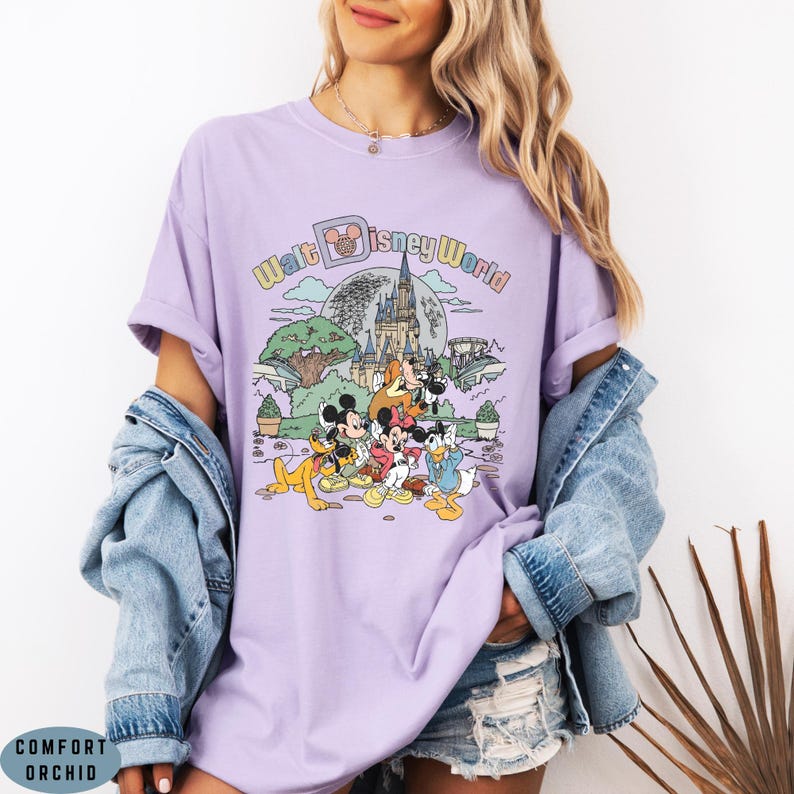 May include: A lilac-colored t-shirt featuring a graphic of Disney characters in front of a castle and the words "Walt Disney World". The design includes Mickey Mouse, Minnie Mouse, Donald Duck, and Goofy. The shirt has short sleeves and a crew neck.