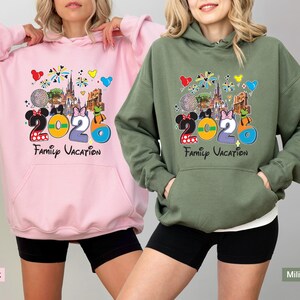 May include: Two hooded sweatshirts, one pink and one olive green, featuring a colorful graphic. The graphic includes the number "2020", Disney characters, and the words "Family Vacation". The sweatshirts have a front pocket and a drawstring hood.