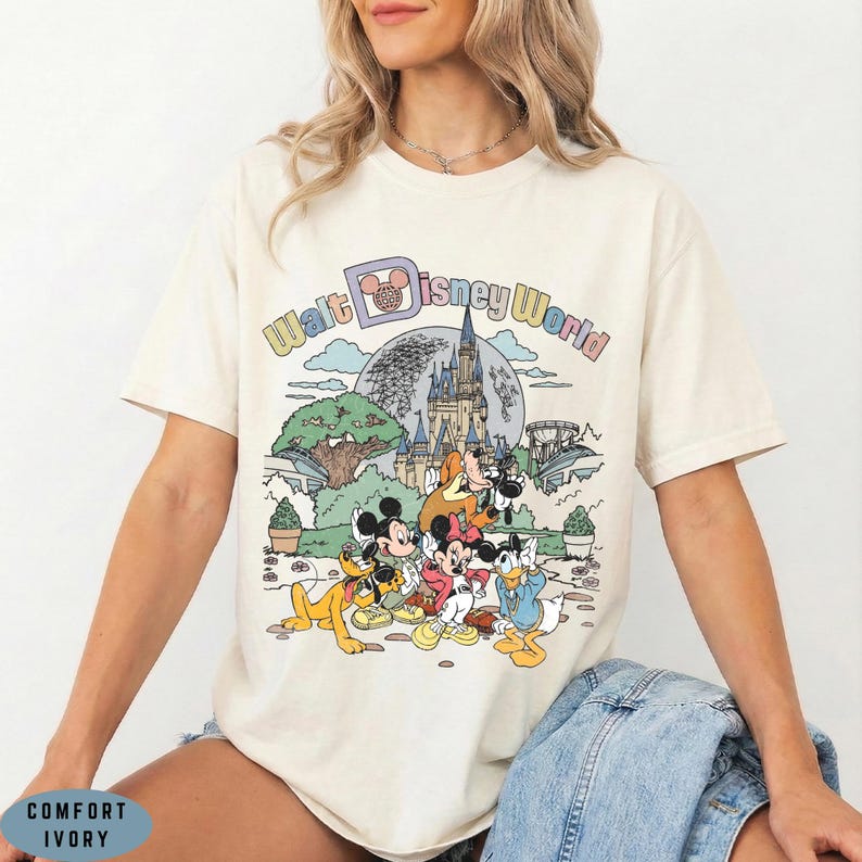 May include: Cream-colored t-shirt featuring a colorful graphic of Disney characters in front of a castle, with the words "Walt Disney World" above. The shirt has short sleeves and a relaxed fit.