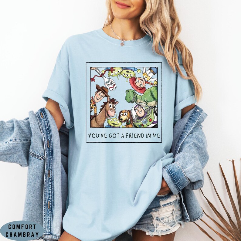 May include: A light blue t-shirt featuring a square graphic print of various animated characters arranged in a circle. Below the characters, the text reads YOU'VE GOT A FRIEND IN ME. The shirt is worn with a denim jacket and denim shorts.