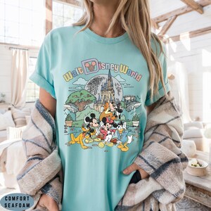 May include: Seafoam green t-shirt featuring a colorful graphic of Disney characters and the text "Walt Disney World." The design includes Mickey Mouse, Minnie Mouse, Donald Duck, and Pluto in front of a castle and a full moon.