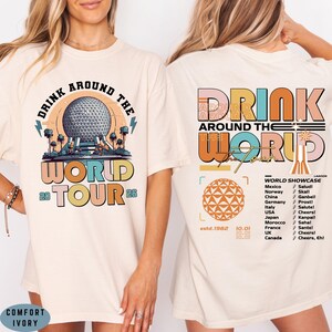 May include: Ivory-colored t-shirt with a colorful graphic design. The front features the text "DRINK AROUND THE WORLD TOUR 26" with a graphic of a sphere and trees. The back has "DRINK AROUND THE WORLD" and a list of countries.