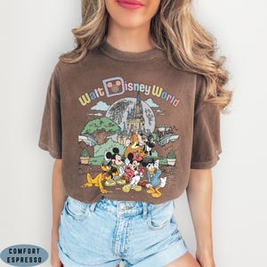 May include: Brown t-shirt with a vintage-style graphic featuring Disney characters and the text "Walt Disney World." The design includes Mickey Mouse, Minnie Mouse, Donald Duck, and Pluto in front of a castle and park scene. The shirt is paired with denim shorts.