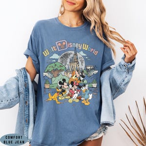 May include: Blue Comfort Color t-shirt featuring a colorful graphic of Disney characters in front of a castle and the words "Walt Disney World". The shirt has rolled-up sleeves and a relaxed fit.