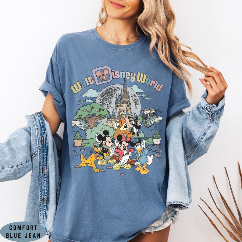 May include: Blue Comfort Color t-shirt featuring a colorful graphic of Disney characters in front of a castle and the words "Walt Disney World". The shirt has rolled-up sleeves and a relaxed fit.