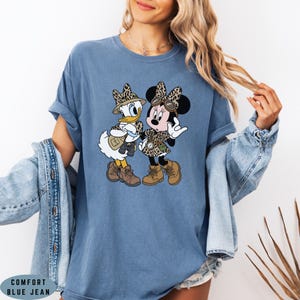 K&ouml;nnte beinhalten: Ein blaues Kurzarm-T-Shirt mit einem Grafikdruck von Daisy Duck und Minnie Mouse in Safari-Outfits mit Leopardenmuster-H&uuml;ten, braunen Stiefeln und Entdeckerausr&uuml;stung. Das Shirt hat einen Rundhalsausschnitt und eine lockere Passform.