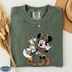 K&ouml;nnte beinhalten: Ein moosgr&uuml;nes T-Shirt mit Rundhalsausschnitt und einem Aufdruck von Daisy Duck und Minnie Mouse in Safari-Kleidung mit Leopardenmuster-Akzenten. Das Shirt hat ein Comfort Colors-Etikett am Kragen und eine silberne Stern-Halskette.