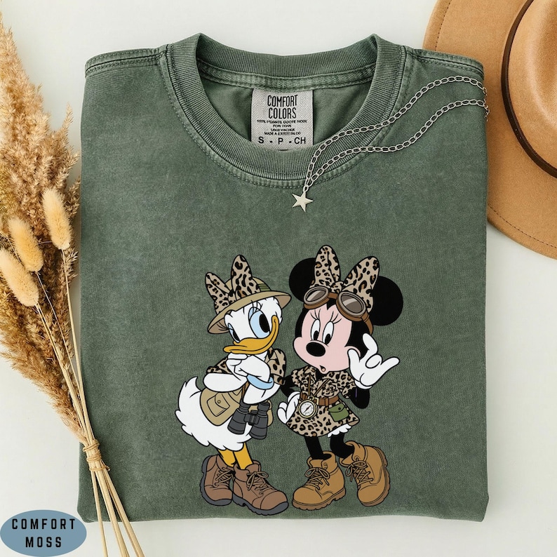 K&ouml;nnte beinhalten: Ein moosgr&uuml;nes T-Shirt mit Rundhalsausschnitt und einem Aufdruck von Daisy Duck und Minnie Mouse in Safari-Kleidung mit Leopardenmuster-Akzenten. Das Shirt hat ein Comfort Colors-Etikett am Kragen und eine silberne Stern-Halskette.