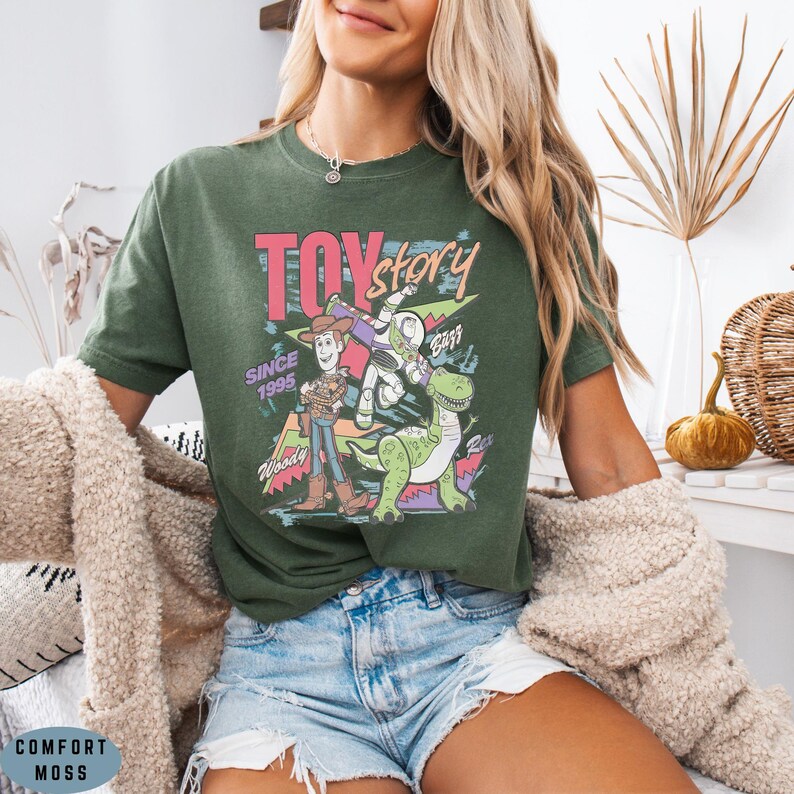 May include: An olive green short-sleeve t-shirt featuring a graphic print of Toy Story characters Woody, Buzz Lightyear, and Rex. The design includes the text TOY story, SINCE 1995, Woody, Buzz, and Rex in colourful, retro-style lettering and shapes.