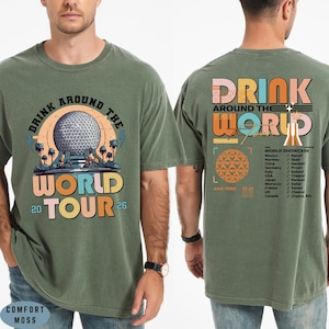 May include: Olive green t-shirt with a graphic design. The front features the text "DRINK AROUND THE WORLD TOUR 20 26" with a colorful illustration. The back has "DRINK AROUND THE WORLD" and a list of countries.