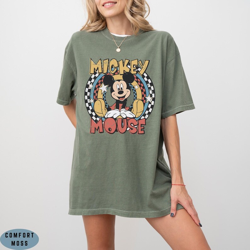 Puede incluir: Una camiseta de manga corta color verde oliva con un gr&aacute;fico de Mickey Mouse centrado en un marco circular. El dise&ntilde;o incluye un borde a cuadros blanco y negro, detalles en amarillo y azul, estrellas blancas y el texto MICKEY MOUSE en rojo.