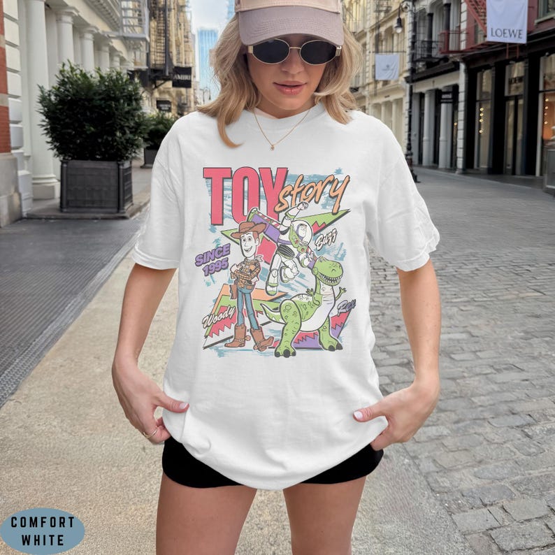 May include: A white short-sleeved t-shirt featuring a colourful graphic print of Toy Story characters Woody, Buzz Lightyear, and Rex. The design includes the text TOY story, SINCE 1995, Woody, Buzz, and Rex in a retro-inspired, multi-coloured layout.
