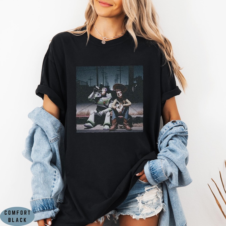 May include: A black crew neck t-shirt featuring a graphic print of the characters Buzz Lightyear and Woody sitting together on a curb at night. The shirt has short sleeves and a relaxed fit, with the text COMFORT BLACK visible in the corner.
